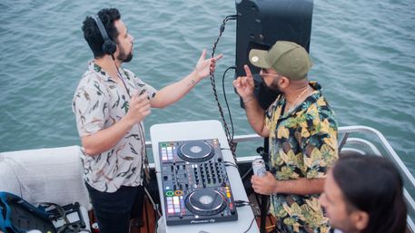 Two DJs performing a lively boat-party DJ set on calm water, mixing on a controller with speakers while wearing tropical shirts.