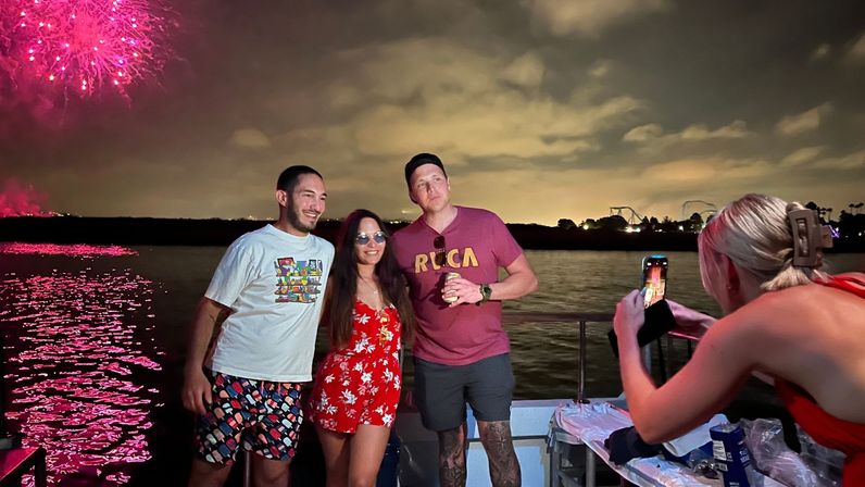 Three people posing on a boat at night as pink fireworks explode over the water, reflecting on the waterfront with distant roller-coaster silhouettes while another person photographs them on a smartphone.