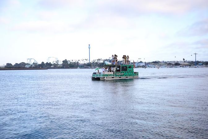 Green party boat with people on the upper deck cruising a calm coastal harbor, palm trees and amusement-park rides on the distant waterfront.