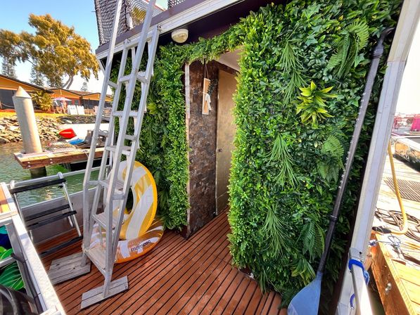 Dockside boat deck at a marina with wooden planks, an aluminum ladder, paddle and inflatable tube, and a lush vertical green wall framing a doorway.