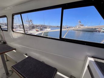 Sunny marina view from a boat cabin: bench seats and tables inside, large windows framing docked yachts, masts, and calm blue water