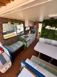 Tropical-themed houseboat interior with compact white galley kitchen, booth dining and cushioned benches, faux green wall with decorative parrot, tiki surfboard sign, wood-style deck flooring and marina view through the window.