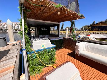 Sunny marina dock with a pontoon-style boat deck under a bamboo canopy, leafy green vines, cushioned bench seating and orange teak-style flooring beside calm waterfront and nearby marina buildings.