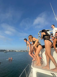 Group of women in bikinis lined along a yacht edge, about to jump into a sunny coastal bay with paddleboarders and harbor buildings in the distance.