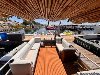 Sunlit marina view from beneath a bamboo canopy on a pontoon boat, showing cushioned seating, wooden deck, colorful jet skis lined on the dock and moored boats on calm water.
