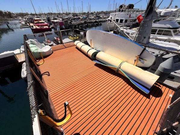 Sunny marina dock with orange slatted deck, rolled foam mats and a paddleboard leaning near moored yachts in a coastal harbor.