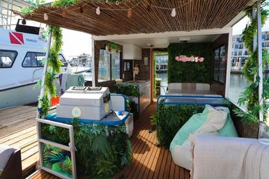 Cozy tropical-themed houseboat lounge docked at a marina with wooden decking, hanging greenery, small kitchenette, cushioned bench and beanbag seating.