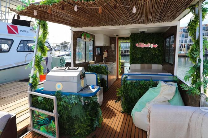Cozy tropical-themed houseboat lounge docked at a marina with wooden decking, hanging greenery, small kitchenette, cushioned bench and beanbag seating.