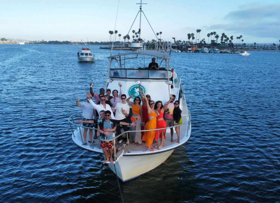 Boat party on a white yacht: cheerful group on the bow waving and holding drinks in a palm-lined coastal harbor with calm blue water.