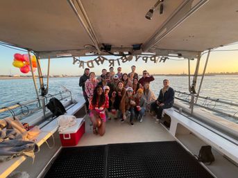 Group of people posing on a covered party boat at sunset with a 'Happy Birthday' banner overhead, colorful balloons tied to the rail, red cups and cooler, and a calm bay and shoreline in the background.