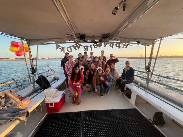 Group of people posing on a covered party boat at sunset with a 'Happy Birthday' banner overhead, colorful balloons tied to the rail, red cups and cooler, and a calm bay and shoreline in the background.