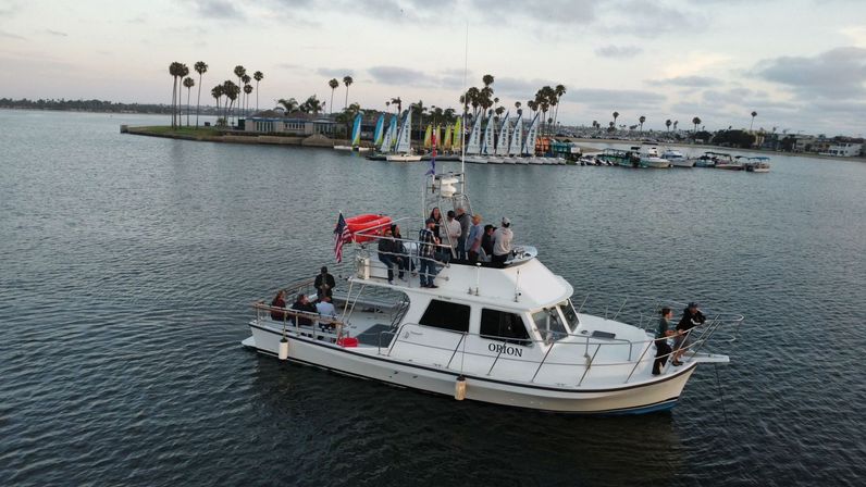 White cabin cruiser with a group of people and an American flag cruising a calm coastal bay past a palm-tree lined marina with colorful sailboats under a cloudy sky.