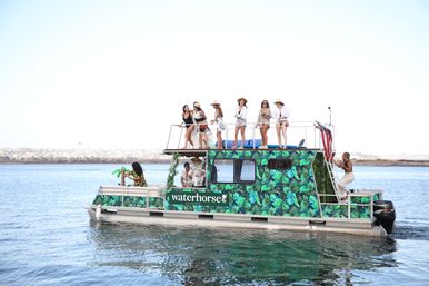 Tropical-patterned two-level pontoon boat on calm coastal waters with a sunlit group on the upper deck enjoying drinks and views during a summertime boat party.