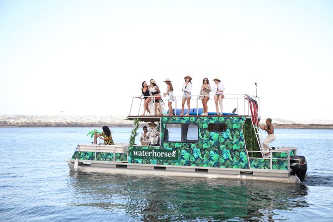 Tropical-patterned two-level pontoon boat on calm coastal waters with a sunlit group on the upper deck enjoying drinks and views during a summertime boat party.