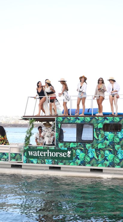 Tropical-patterned two-level pontoon boat on calm coastal waters with a sunlit group on the upper deck enjoying drinks and views during a summertime boat party.