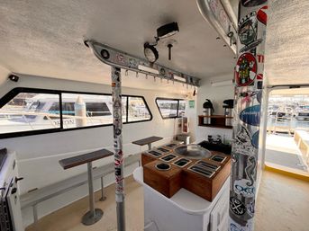 Sunlit fishing boat cabin at a marina with a wooden bait/tackle station of metal bins, sticker-covered support poles, bench seating and windows showing docked boats