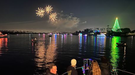 Nighttime holiday lighted boat parade on a calm bay — decorated boats and colorful reflections, a giant green illuminated Christmas tree on shore, fireworks bursting overhead and spectators watching from a boat.
