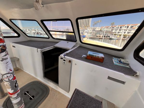 Sunlit boat cabin interior with carpeted storage benches, an open hatch, and large windows framing a marina with docked boats, palm trees and waterfront buildings.
