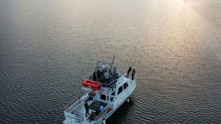 Aerial view of a white motor yacht with a group of people on deck and an American flag, drifting on calm rippled water at sunset — evening coastal scene