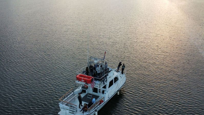 Aerial view of a white motor yacht with a group of people on deck and an American flag, drifting on calm rippled water at sunset — evening coastal scene