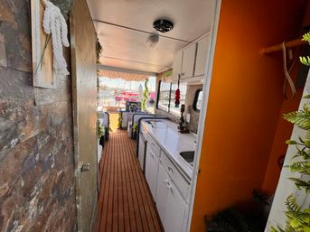 Cozy houseboat interior with narrow teak walkway, white galley kitchen and orange accent wall opening to a marina view with boats
