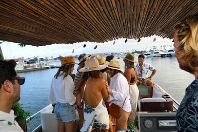 Group in summer hats socializing under a bamboo canopy on a pontoon boat at a marina with yachts and palm trees