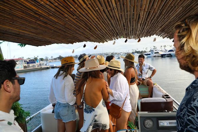 Group in summer hats socializing under a bamboo canopy on a pontoon boat at a marina with yachts and palm trees