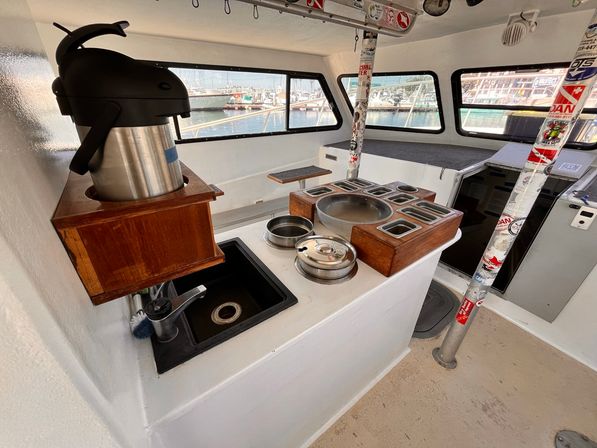 Cozy boat galley interior with stainless airpot coffee dispenser on a wooden shelf, small black sink and countertop holding stacked pots and a wooden spice/cup organizer, with large windows looking out to a marina of docked boats.