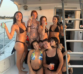 Seven friends in bikinis laughing and holding drinks on a sunlit boat deck during a coastal sunset cruise.