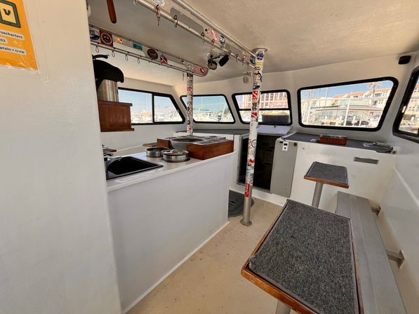 Bright white motorboat cabin with a compact galley, sink and cookware, carpeted bench seats, sticker-covered support poles and large windows framing a sunny marina with docked boats.