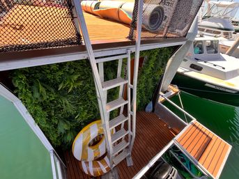 Marina floating deck with metal ladder to an upper platform, faux vertical green wall, two yellow-and-white swim rings, and a nearby motorboat on green water.