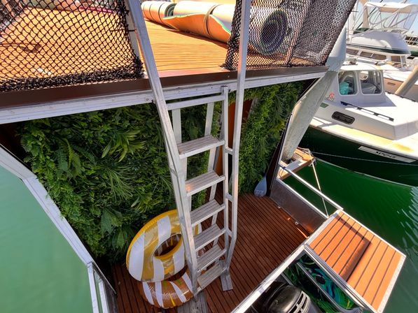 Marina floating deck with metal ladder to an upper platform, faux vertical green wall, two yellow-and-white swim rings, and a nearby motorboat on green water.