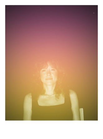 Seated woman portrait with a gentle smile against a purple-to-gold gradient light-leak backdrop, vintage film-style effect, wearing a black top and delicate necklace.