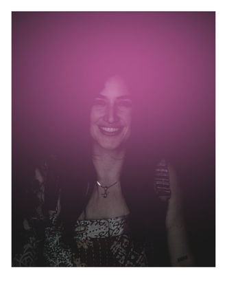 Low-light portrait of a smiling woman from chest up with dark hair, patterned top and pendant necklace, soft magenta light overlay
