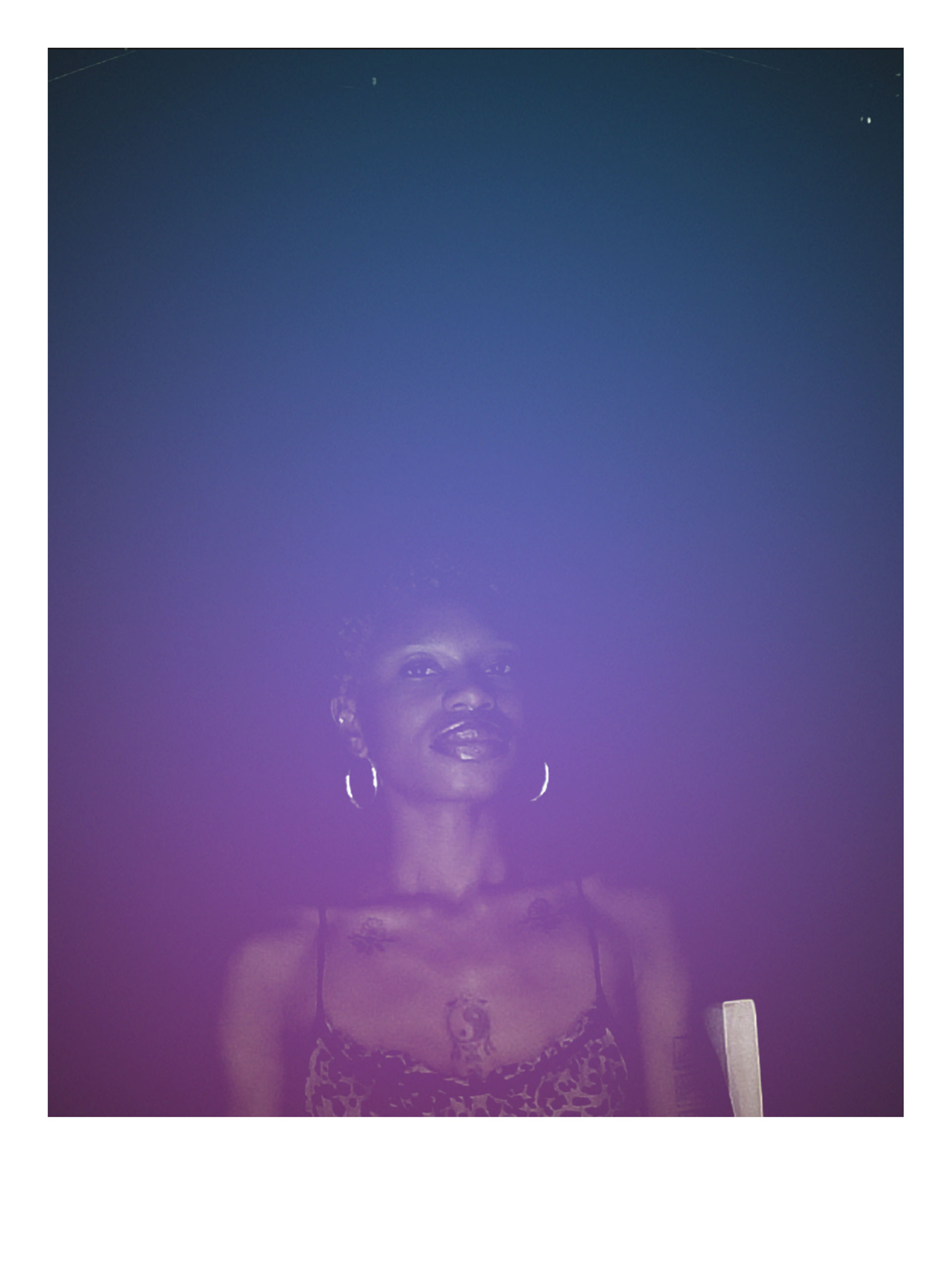 Moody night portrait of a woman bathed in purple-blue neon light, wearing hoop earrings and a patterned spaghetti-strap top with visible chest tattoos against a dark background.