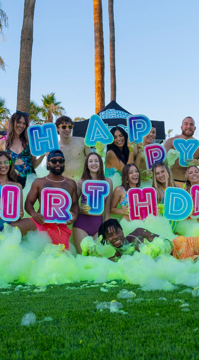 Smiling group of young adults at a sunny outdoor foam party by palm trees, in swimwear holding colorful "HAPPY BIRTHDAY" letter signs amid neon green foam on grass