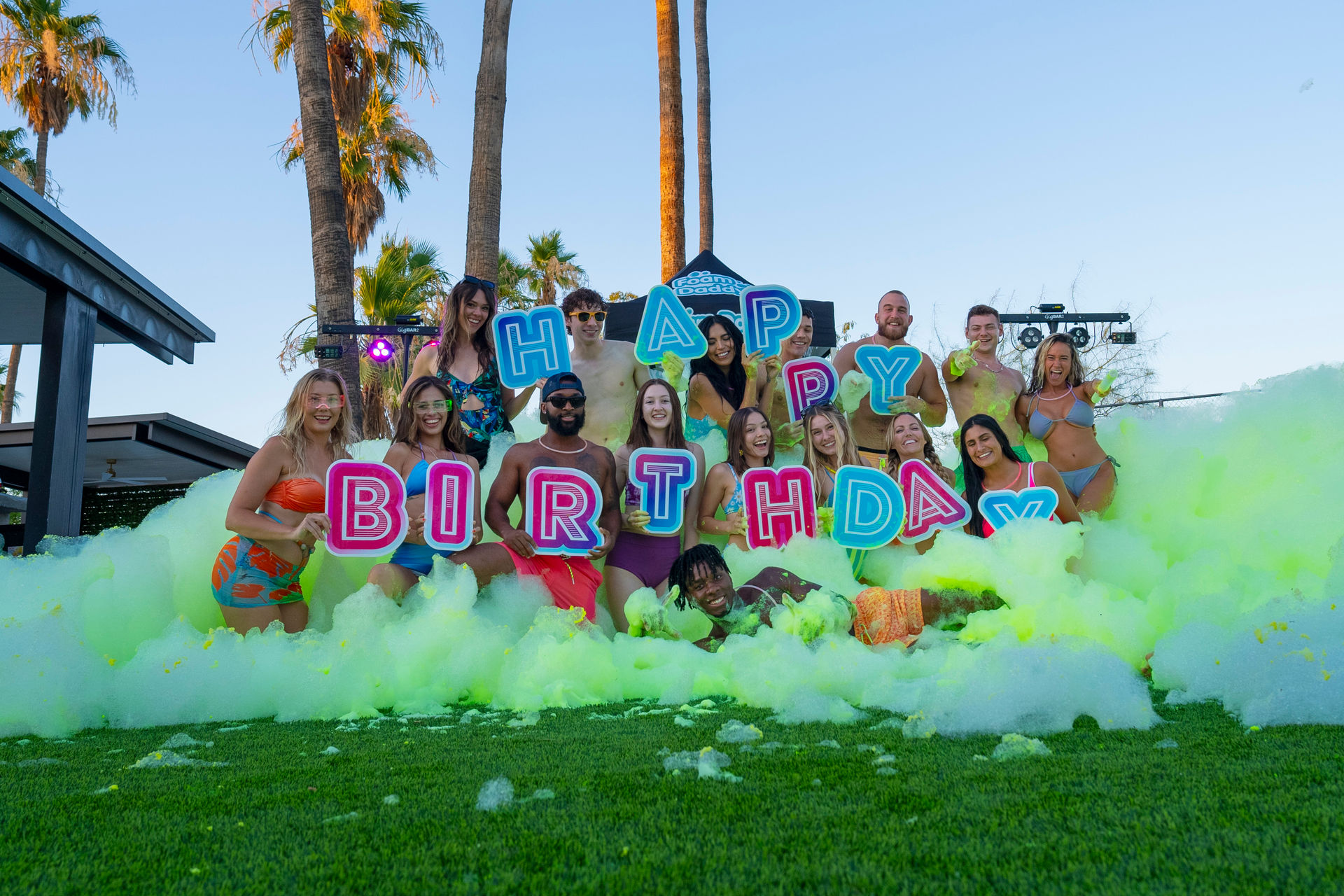 Smiling group of young adults at a sunny outdoor foam party by palm trees, in swimwear holding colorful "HAPPY BIRTHDAY" letter signs amid neon green foam on grass