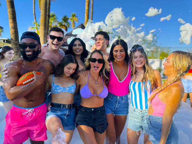 Smiling group of friends at an outdoor foam party with palm trees, colorful swimwear, soap bubbles and an orange football.