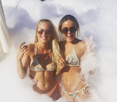 Two people in sequined bikinis and sunglasses laughing amid thick foam at a sunny outdoor pool foam party, playful summer vibes