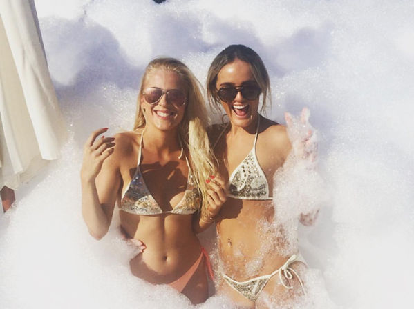 Two women in sparkly bikinis and sunglasses laughing amid white bubbles at a sunny outdoor foam party, playful summer pool party vibe.