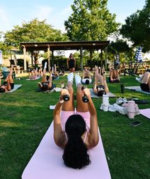 Sunny outdoor group fitness class on a park lawn — participants on yoga mats doing leg raises with dumbbells while an instructor leads from under a wooden pergola, surrounded by trees and picnic benches.