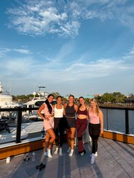 Five women in colorful workout clothes smiling arm-in-arm on a marina boardwalk at sunset with yachts and calm water behind them, blue sky with wispy clouds, a yoga mat, dumbbells and water bottles on the deck