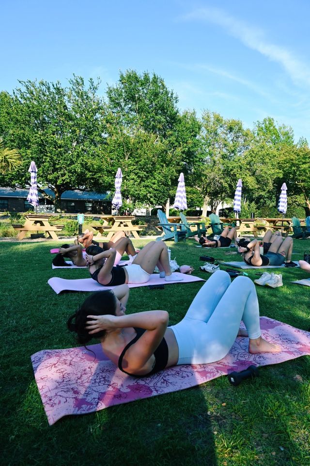 Outdoor group fitness class with people doing crunches on yoga mats on a sunny grassy lawn, picnic tables and striped umbrellas, leafy trees in the background