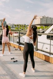 Two women doing a sunny outdoor stretch/yoga workout on mats at a marina dock with boats and waterfront condos in the background.