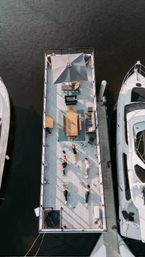 Aerial view of a sunlit floating deck docked in a marina between yachts, featuring a patio umbrella, dining table, lounge seating and a small group doing an outdoor workout on the waterfront.