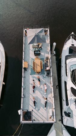 Aerial view of a sunlit floating deck docked in a marina between yachts, featuring a patio umbrella, dining table, lounge seating and a small group doing an outdoor workout on the waterfront.