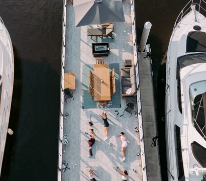 Aerial view of a sunlit floating deck docked in a marina between yachts, featuring a patio umbrella, dining table, lounge seating and a small group doing an outdoor workout on the waterfront.
