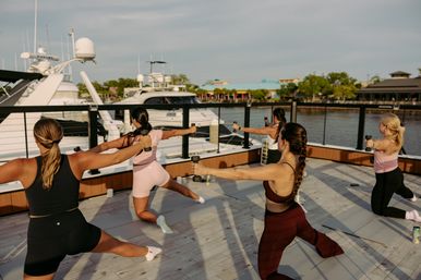 Group of women powering through a waterfront dumbbell workout on a marina deck with yachts and calm water in the background.