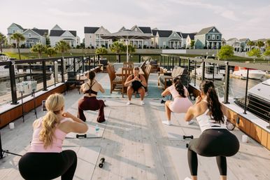 Women doing a group outdoor workout on a sunlit marina deck — instructor leads squats on yoga mats with dumbbells, boats docked nearby and pastel coastal houses across the water.