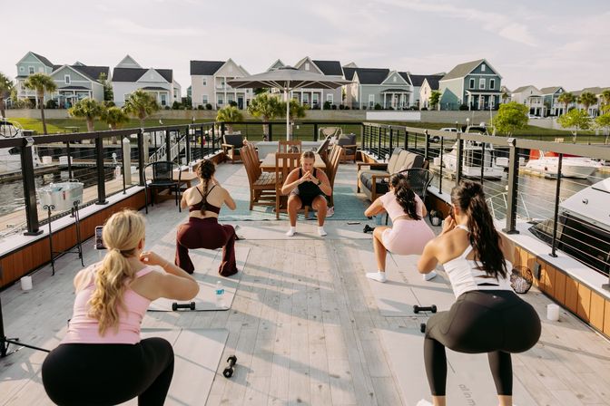 Women doing a group outdoor workout on a sunlit marina deck — instructor leads squats on yoga mats with dumbbells, boats docked nearby and pastel coastal houses across the water.